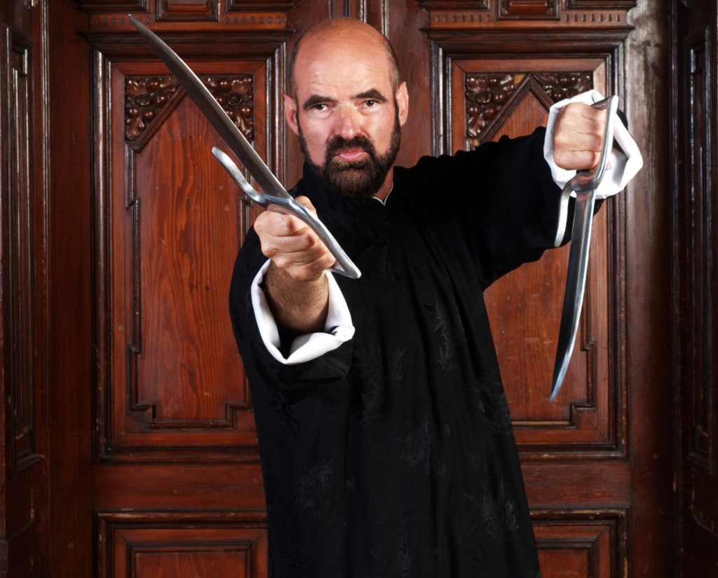 Professor Doctor Keith R. Kernspecht in a traditional black WingTsun outfit, pointing two sabers forward and facing the camera head-on.